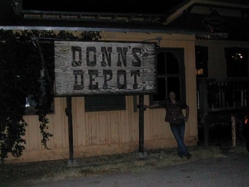 Nighttime exterior of Donn’s Depot in Austin, with a person leaning against the large, rustic wooden sign at the entrance to the depot-style building.