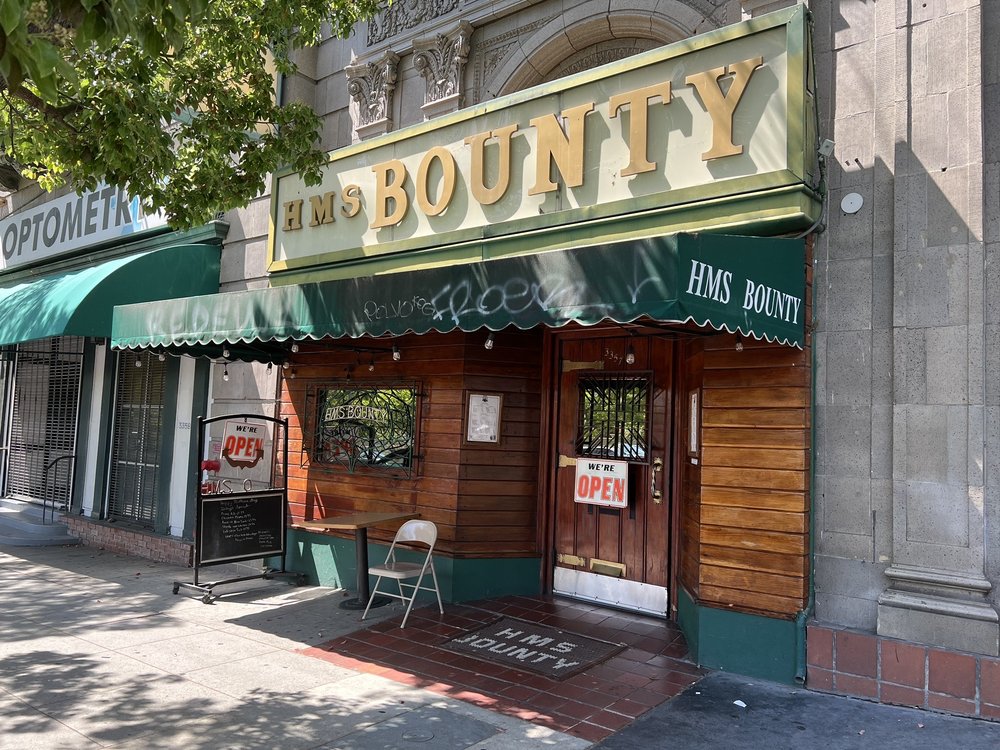 Street-level exterior of HMS Bounty in Los Angeles, a classic dive bar with a "WE'RE OPEN" sign, wood-paneled entrance, and green awnings. A top result for 'best dive bars near me' in LA.