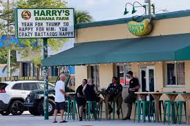 Outside view of Harry's Banana Farm in Lake Worth, Florida, a casual dive bar with patrons sitting at high-top tables under a green awning.