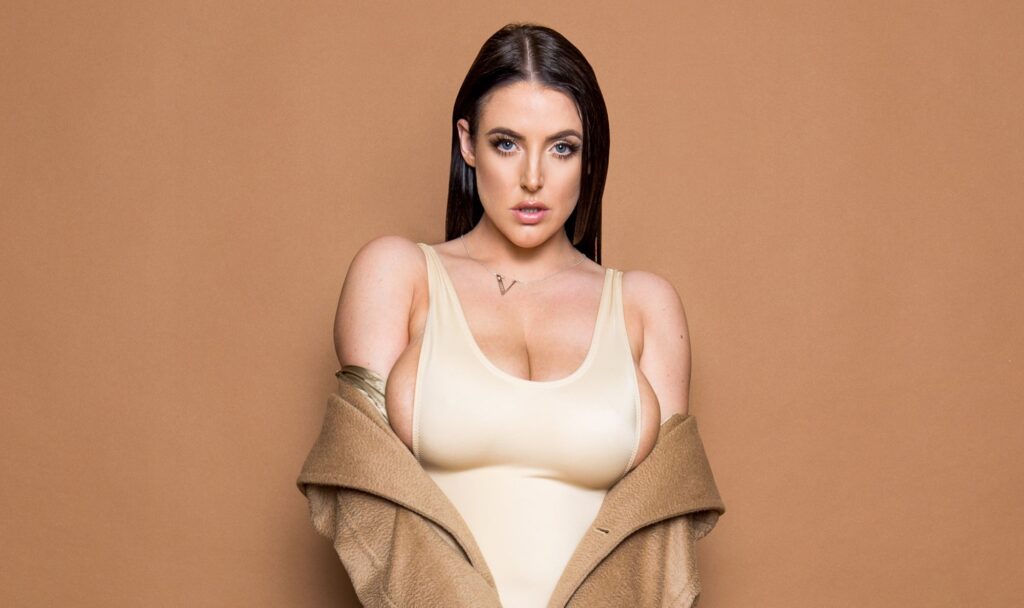 A headshot of Angela White, an adult film actress, with long dark hair, blue eyes, and a light top under a brown coat against a beige background, related to Angela White leaks.