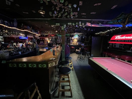 Dimly lit interior of Mother Lode Bar & Deli in Sacramento, featuring a long wooden bar, a red pool table, and dollar bills covering the ceiling. A great example of the 'best dive bars near me' in Sacramento.