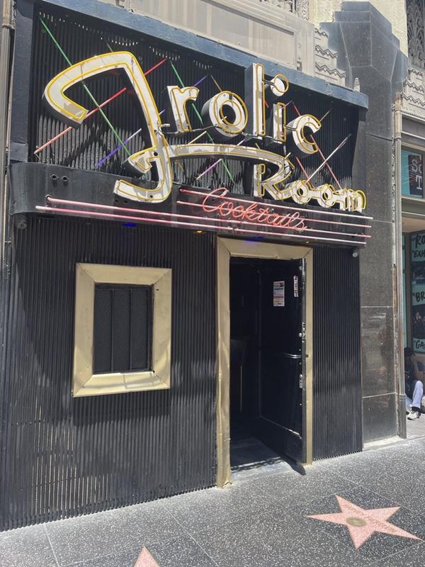 Exterior of The Frolic Room in Los Angeles, featuring its iconic, retro neon sign, a black corrugated facade, and a Hollywood Walk of Fame star on the sidewalk.