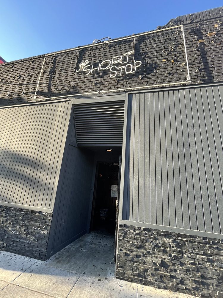 Exterior of The Short Stop, a Los Angeles dive bar, showing a grey facade, brick upper wall with a white neon sign, and an open doorway. A classic spot when searching for the 'best dive bars near me'
