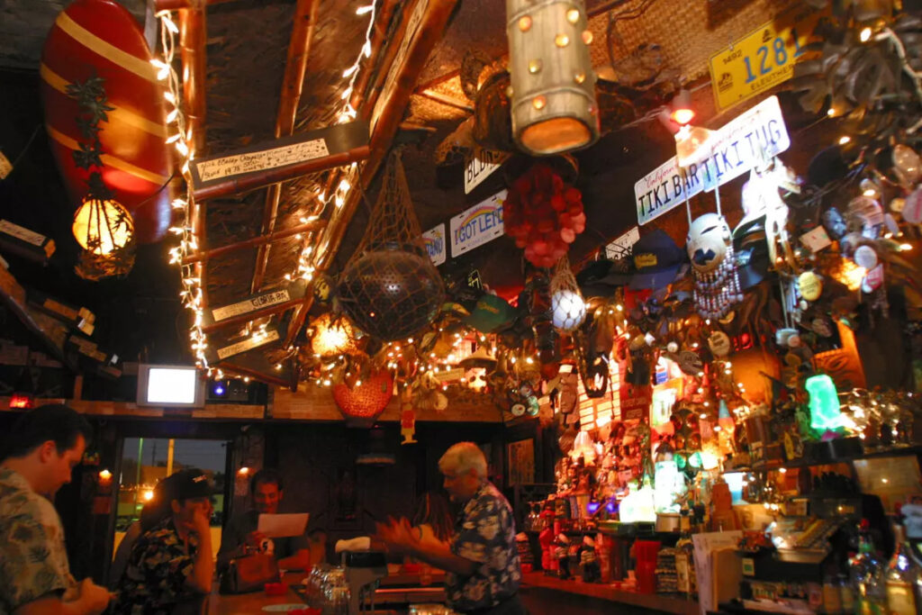 The vibrant and densely decorated interior of Tiki Ti in Los Angeles, packed with patrons at the bar and surrounded by tiki masks, string lights, and eclectic tropical decor.