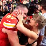 Travis Kelce in his red Chiefs jersey embracing a woman (likely his girlfriend or a fan) amid a crowd of photographers and people, following a milestone or a Travis Kelce Touchdown Dance.