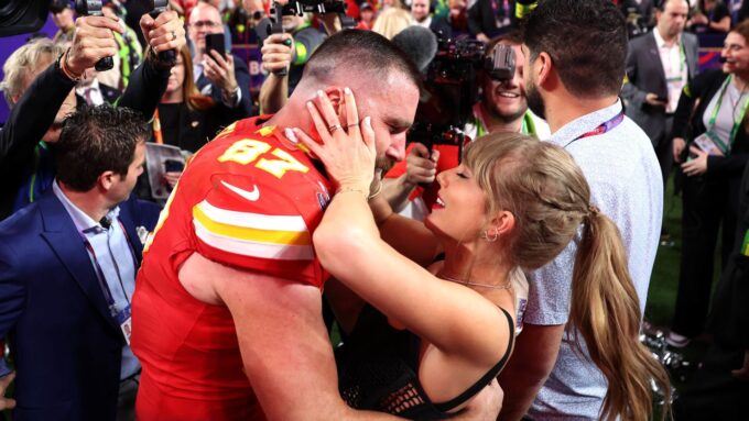 Travis Kelce in his red Chiefs jersey embracing a woman (likely his girlfriend or a fan) amid a crowd of photographers and people, following a milestone or a Travis Kelce Touchdown Dance.