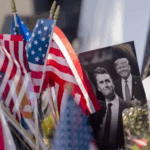 A close-up photograph of small American flags planted in the ground, surrounding a picture of two men, one smiling and giving a thumbs up, with a second, smaller picture of a smiling Donald Trump and Charlie Kirk overlaid in the upper right corner emphasizes the issue that why Trump Cancels Student Visas.