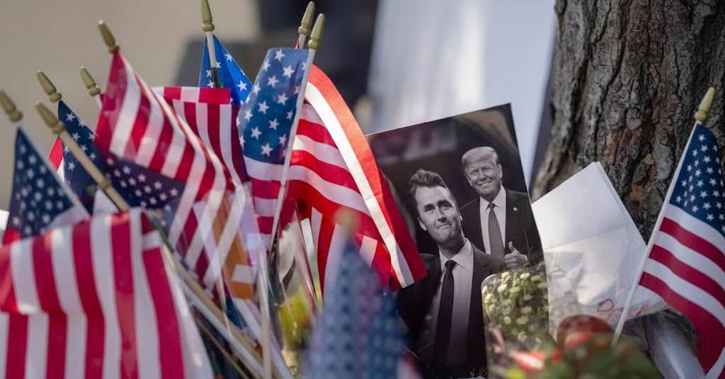 A close-up photograph of small American flags planted in the ground, surrounding a picture of two men, one smiling and giving a thumbs up, with a second, smaller picture of a smiling Donald Trump and Charlie Kirk overlaid in the upper right corner emphasizes the issue that why Trump Cancels Student Visas.