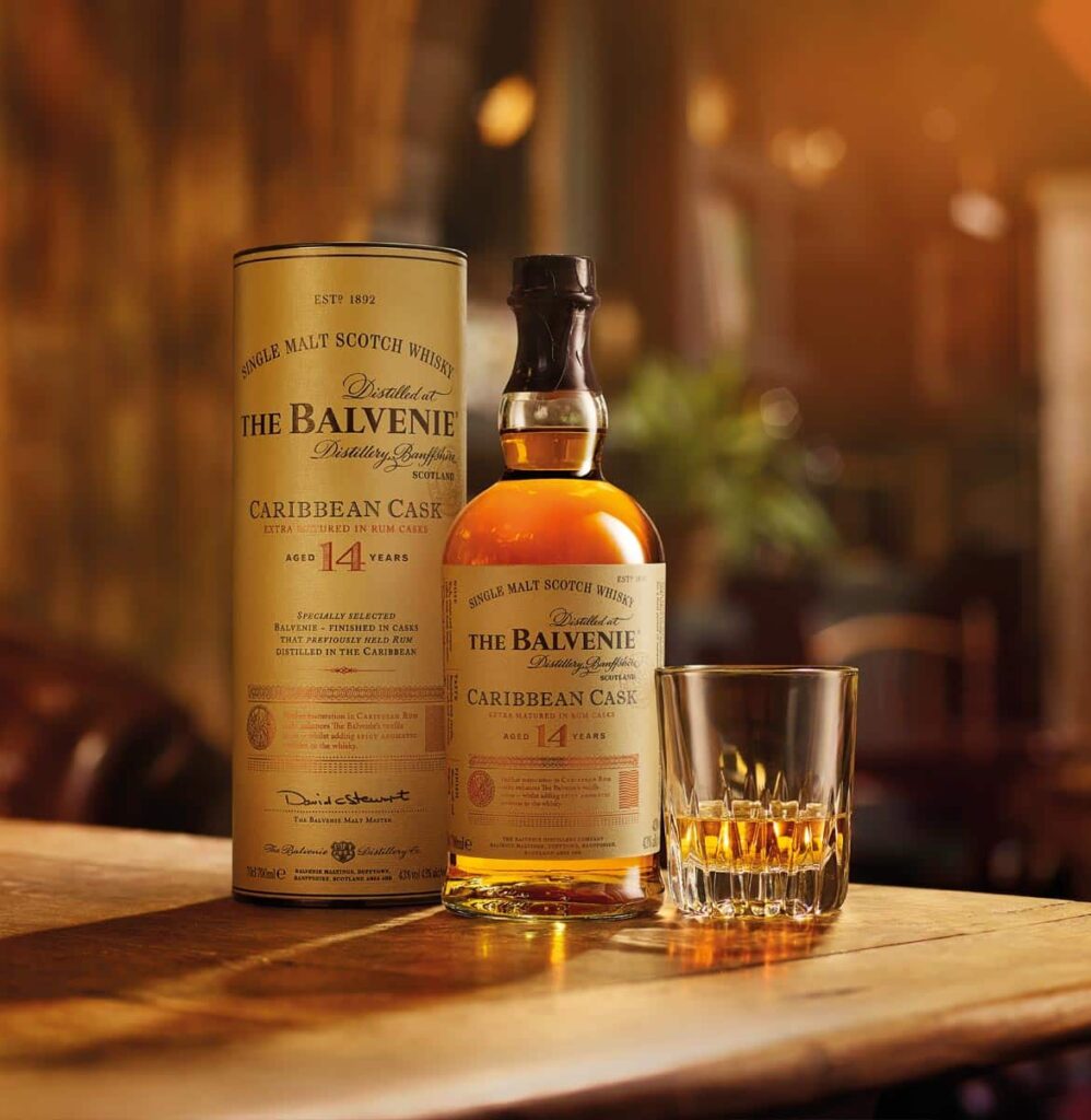 A bottle of The Balvenie 14 Year Old Caribbean Cask Single Malt Scotch Whisky next to a full glass of whiskey on a wooden table. A tall, cylindrical cardboard tube container for the bottle is visible behind it.