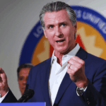 A color photograph of California Governor Gavin Newsom speaking at a podium during a Prop 50 event, raising both fists in a gesture of triumph or emphasis.