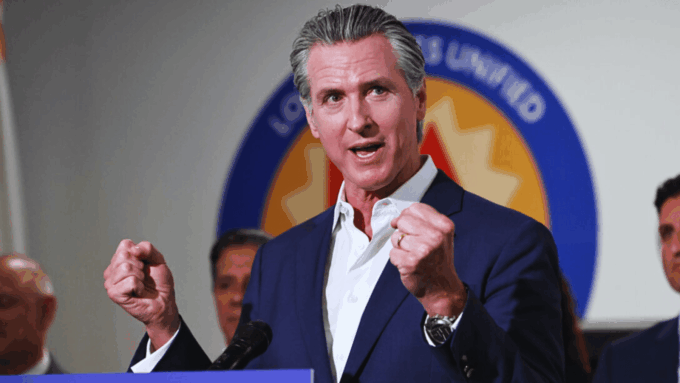 A color photograph of California Governor Gavin Newsom speaking at a podium during a Prop 50 event, raising both fists in a gesture of triumph or emphasis.