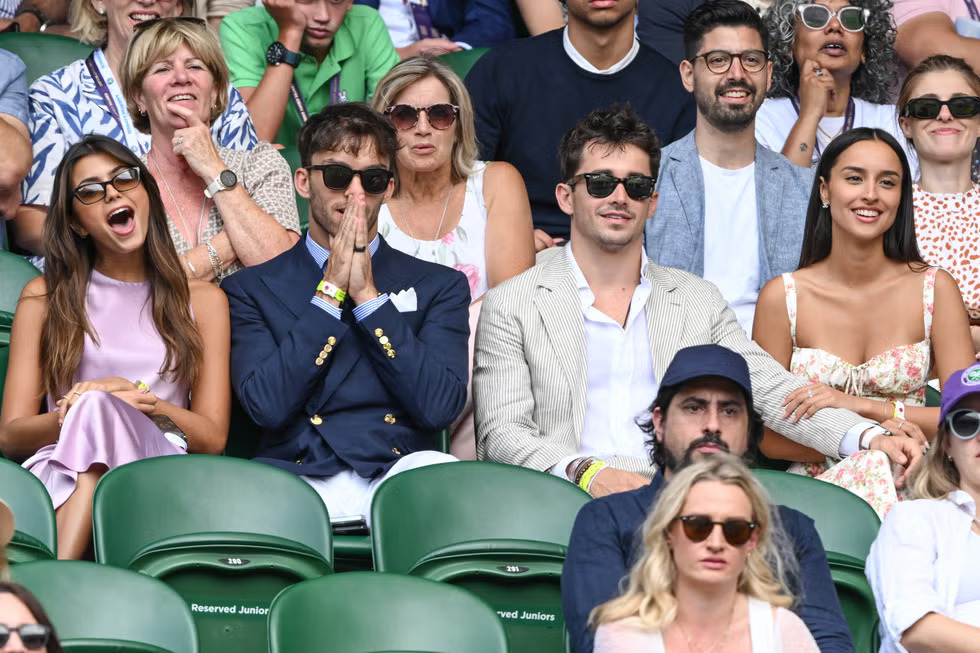Charles Leclerc and Alexandra Saint Mleux sitting in the crowd at Wimbledon, both wearing sunglasses, with other spectators around them.