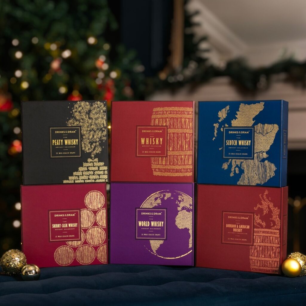Six square, premium-looking advent calendar boxes from "Drinks by the Dram" stacked in two rows of three, against a festive background with a blurred Christmas tree. The boxes are labeled: "Peaty Whisky," "Whisky," "Scotch Whisky," "Sherry Cask Whisky," "World Whisky," and "Bourbon & American Whiskey." Perfect whiskey gift for Advent Calendar 2025