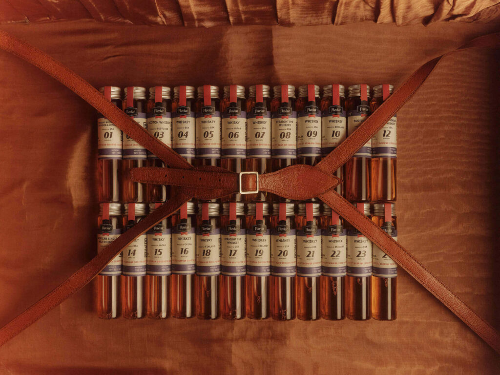 An overhead view of the Flaviar Whiskey Advent Calendar, which is presented in a brown leather-look suitcase box. It contains 24 glass vials (whiskey samples) secured by a central brown leather strap with a buckle. The vials are individually numbered from 1 to 24. Perfect whiskey gift for Advent Calendar 2025.
