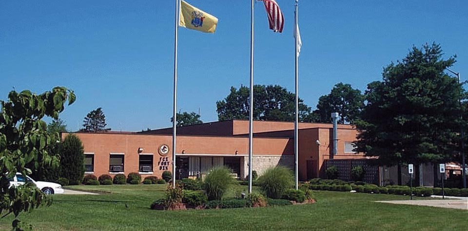 An exterior view of the Fort Dix Federal Correctional Institution building on a sunny day, with three flagpoles flying flags in front of a well-maintained lawn.