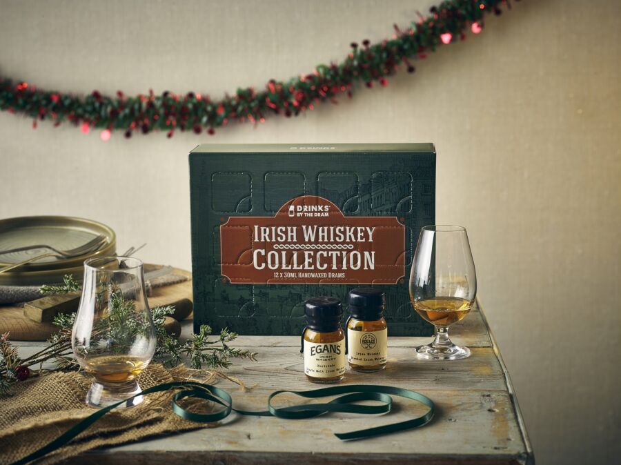 A green "Irish Whiskey Collection" Advent Calendar box by "Drinks by the Dram" placed on a rustic wooden table with two small bottles of whiskey, decorative greenery, and an empty whiskey glass.