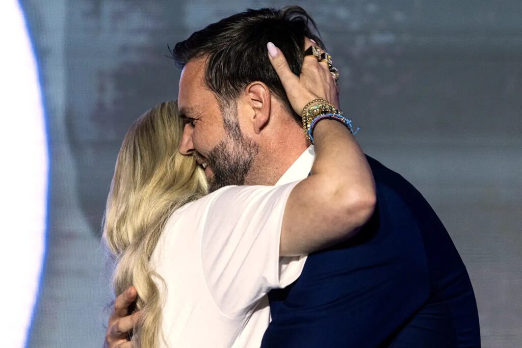 A close-up side-view photo of JD Vance embracing Erika Kirk on stage. Vance, smiling, is wearing a dark suit jacket and a white shirt. Kirk, seen from the back, has long blonde hair, and her hand, adorned with bracelets and rings, is resting on the back of Vance's head.