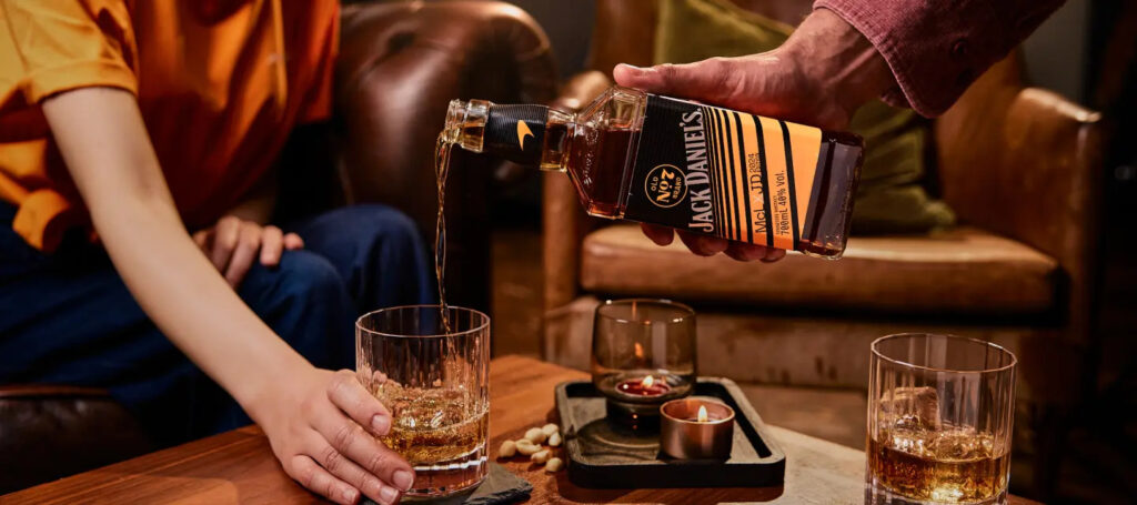 A close-up shot of a person pouring Jack Daniel's Tennessee Whiskey from a bottle into a tall, clear glass being held by another person, while seated in a cozy, dimly lit interior setting.
