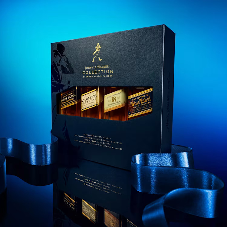 A dark, elegant gift box set from the Johnnie Walker Collection, showcasing four different small bottles: Black Label, Gold Label Reserve, 18 Year Old, and Blue Label, set against a reflective surface and a bright blue background.