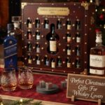 A shot of a large, wooden trunk-style Advent Calendar 2025 for whiskey, surrounded by full-sized bottles, whiskey glasses, and Christmas decorations in front of a fireplace