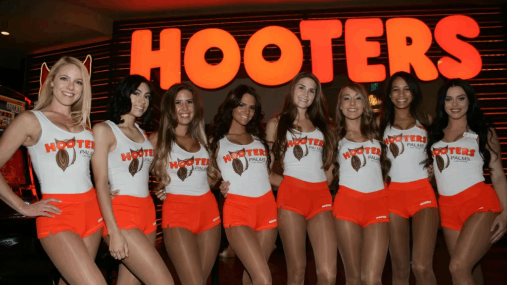 A group of eight girls standing in a row in a warmly lit interior space, all wearing the classic white tank top with the Hooters logo, orange shorts, and sheer pantyhose. A large, illuminated "HOOTERS" sign is visible in the background.