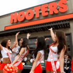 A group of several Hooters girls outside the restaurant building, raising their arms and celebrating. They are wearing sashes over their uniforms (white tops and orange shorts) with the large red "HOOTERS" sign visible above them.