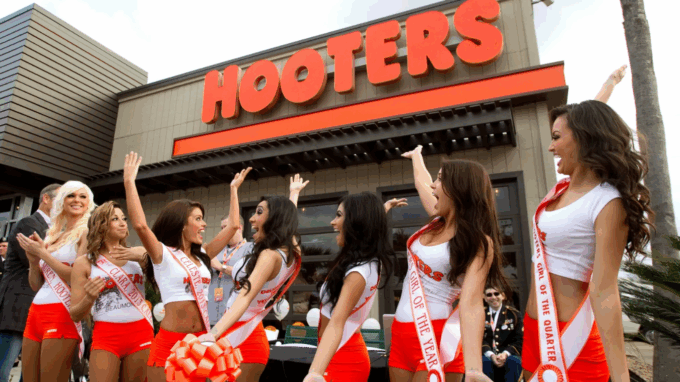 A group of several Hooters girls outside the restaurant building, raising their arms and celebrating. They are wearing sashes over their uniforms (white tops and orange shorts) with the large red "HOOTERS" sign visible above them.