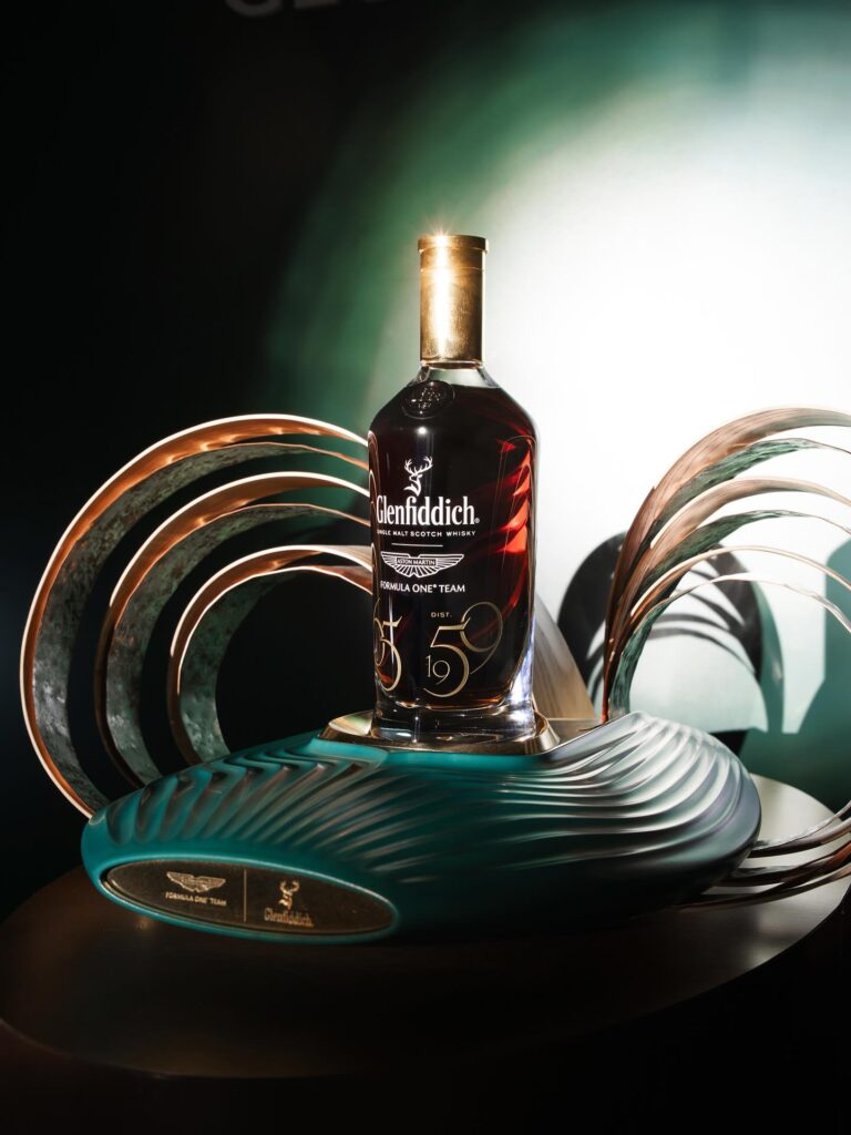 A bottle of Glenfiddich single malt Scotch whisky, displayed on a custom-designed, futuristic-looking teal and bronze stand, possibly part of a collaboration with the Aston Martin Formula 1 team, as indicated by the logo on the stand and bottle label. Perfect whiskey gift for Advent Calendar 2025