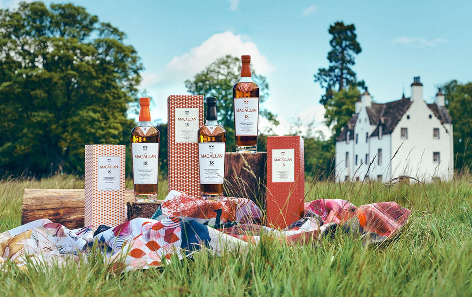 Three bottles of The Macallan 18-Year-Old Single Malt Scotch Whisky and matching rectangular boxes, arranged outdoors on wooden stumps and a colorful fabric scarf in a grassy field, with a traditional white stone building in the background. Perfect whiskey gift for Advent Calendar 2025