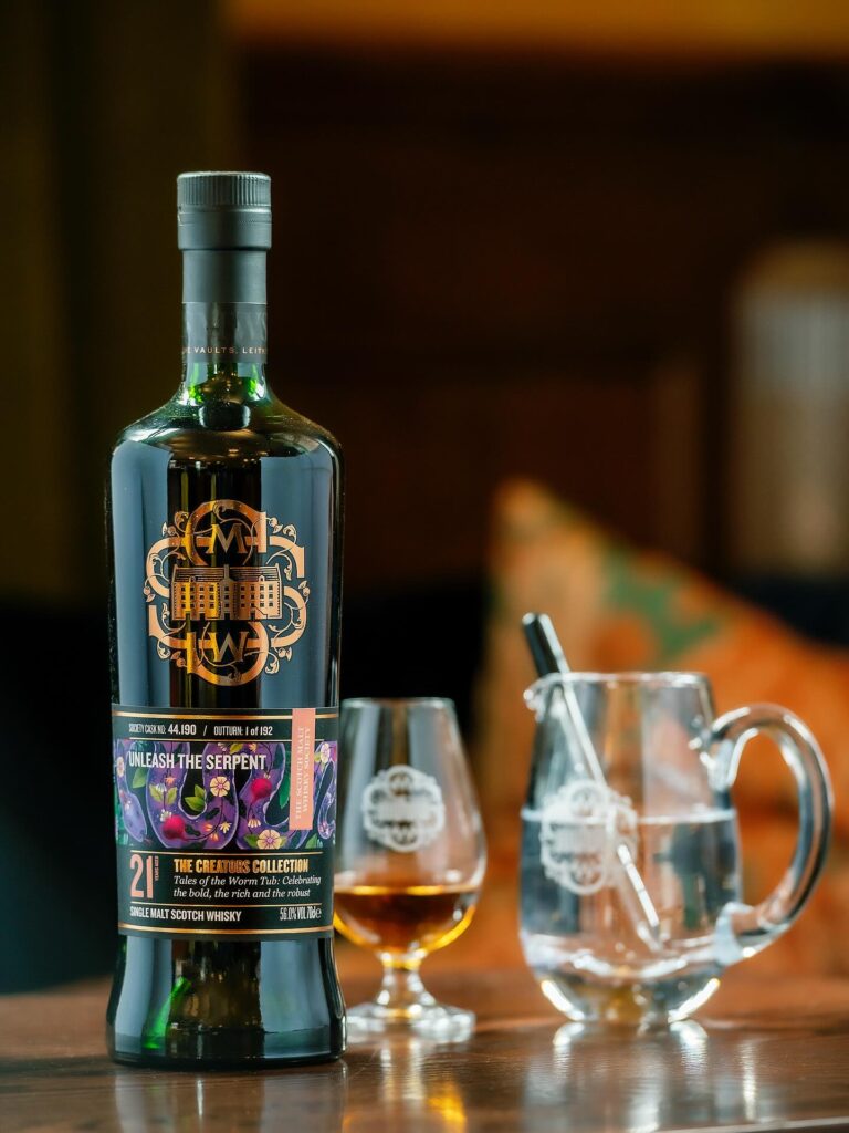 A bottle of Scotch Malt Whisky Society (SMWS) single malt Scotch whisky, labeled "UNLEASH THE SERPENT," standing next to a glass of whisky and a water jug, in a warm, indoor setting.
