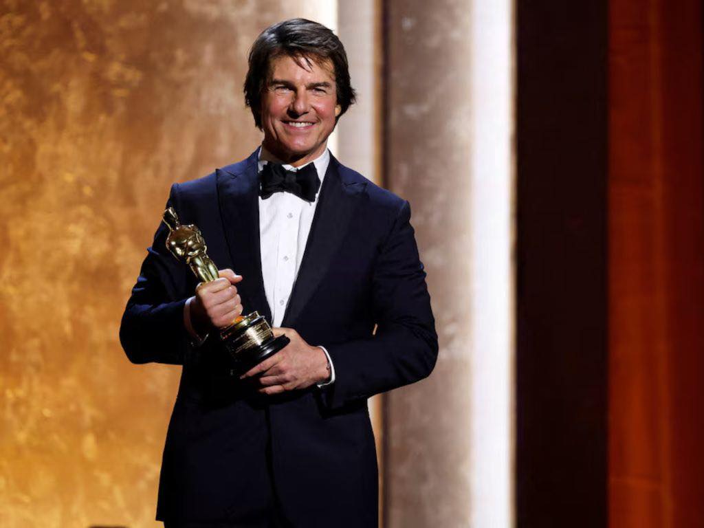 Tom Cruise smiling and holding an Oscar statuette while wearing a tuxedo on stage at an awards ceremony.