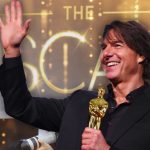 Tom Cruise smiling, waving, and holding an Oscar statuette amidst a crowd of photographers.