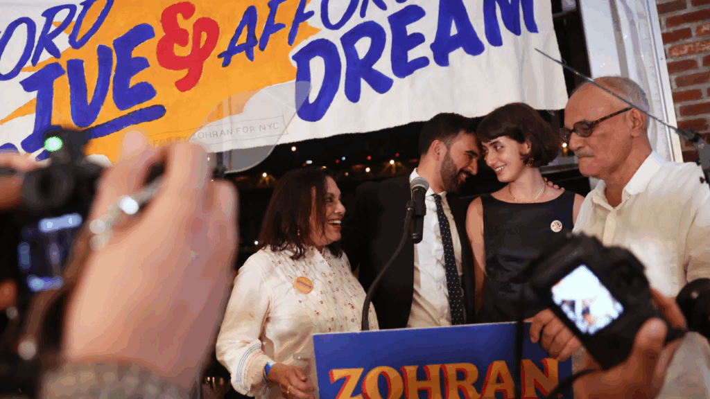 A close-up portrait of Zohran Mamdani, the new Zohran Mamdani NYC Mayor, smiling and wearing a blue blazer, with text referencing his South Asian heritage and family ties.