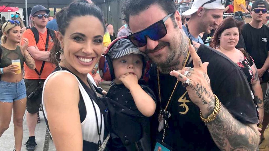 A medium shot of a smiling Bam Margera and Nicole Boyd at an outdoor event. Nicole is on the left with her hair tied back, wearing a black and white top. Bam is on the right, wearing blue-tinted sunglasses, numerous gold chains, and displaying prominent hand tattoos while making a hand gesture. Between them, they hold their young son, Phoenix Wolf, who is wearing a striped hat and red noise-canceling headphones.