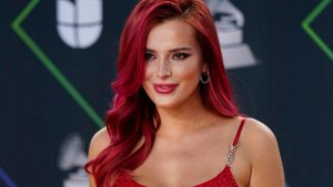 Close-up portrait of Bella Thorne with vibrant, wavy red hair and pink lipstick, smiling slightly while wearing a sparkly red strap dress at a red carpet event.
