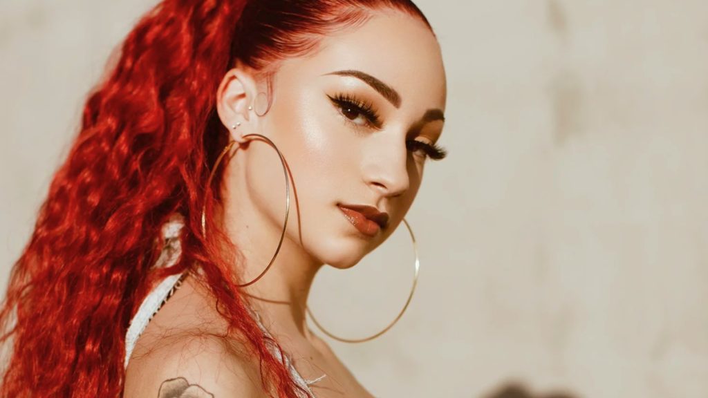 A close-up, high-glamour portrait of Bhad Bhabie looking over her shoulder toward the camera. She has long, vibrant wavy red hair tied in a high ponytail, dramatic winged eyeliner, and very large gold hoop earrings. The lighting is warm and bright against a neutral, sunlit wall.
