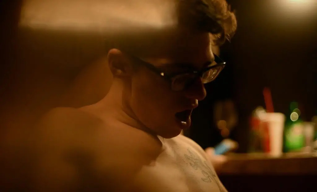 A warm-toned, candid photograph of Lane Rogers wearing glasses in a dimly lit setting. The image shows him from the side with an open-mouthed expression, capturing a moment from his life as a viral creator.