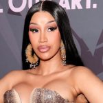 A close-up, chest-up portrait of Cardi B at a red carpet event. She has long, sleek black hair parted down the middle and is wearing large, textured hoop earrings adorned with small silver charms. Her makeup features dramatic lashes and a neutral lip. She is wearing a metallic, bronze-toned structured bodice, set against a blurred background with the word "MUSIC" visible.