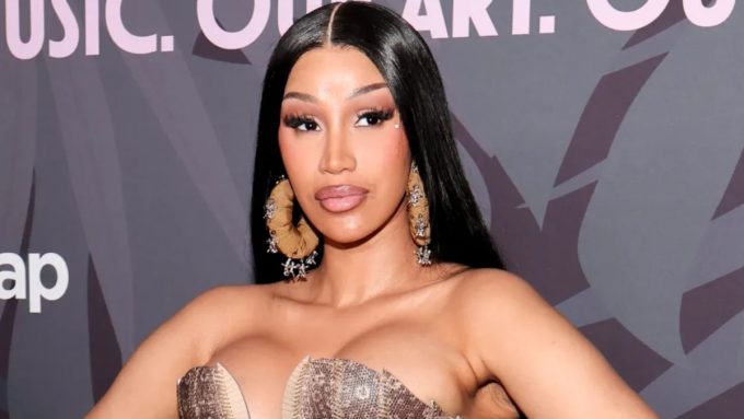 A close-up, chest-up portrait of Cardi B at a red carpet event. She has long, sleek black hair parted down the middle and is wearing large, textured hoop earrings adorned with small silver charms. Her makeup features dramatic lashes and a neutral lip. She is wearing a metallic, bronze-toned structured bodice, set against a blurred background with the word "MUSIC" visible.
