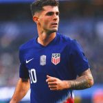 A medium shot of Christian Pulisic during a soccer match. He is wearing a dark blue USA National Team jersey with the number 10 and a captain's patch. He has a focused expression, looking off to the side while running on the pitch. The background shows a blurred stadium crowd.