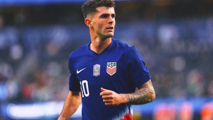 A medium shot of Christian Pulisic during a soccer match. He is wearing a dark blue USA National Team jersey with the number 10 and a captain's patch. He has a focused expression, looking off to the side while running on the pitch. The background shows a blurred stadium crowd.