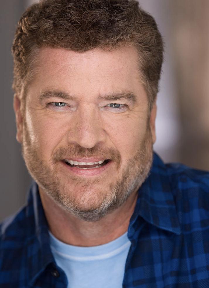 A close-up professional headshot of Pat Finn. He is smiling warmly at the camera, showing a friendly expression. He has short, curly brown hair and a light beard, and he is wearing a blue plaid button-down shirt over a light blue t-shirt.