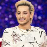 A medium shot of Frankie Grande sitting on a white sofa against a blurred purple and blue background. He is wearing a cream-colored sweater featuring large black-and-white star patterns with red accents and several beaded necklaces. He has blonde, swept-back hair and a groomed beard, looking off-camera with a slight smile.