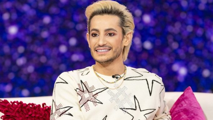 A medium shot of Frankie Grande sitting on a white sofa against a blurred purple and blue background. He is wearing a cream-colored sweater featuring large black-and-white star patterns with red accents and several beaded necklaces. He has blonde, swept-back hair and a groomed beard, looking off-camera with a slight smile.