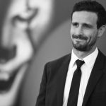 A black and white portrait of actor James Ransone smiling slightly while looking off-camera. He is wearing a dark suit, white shirt, and dark tie. In the blurred background, a large graphic of a clown face is partially visible.