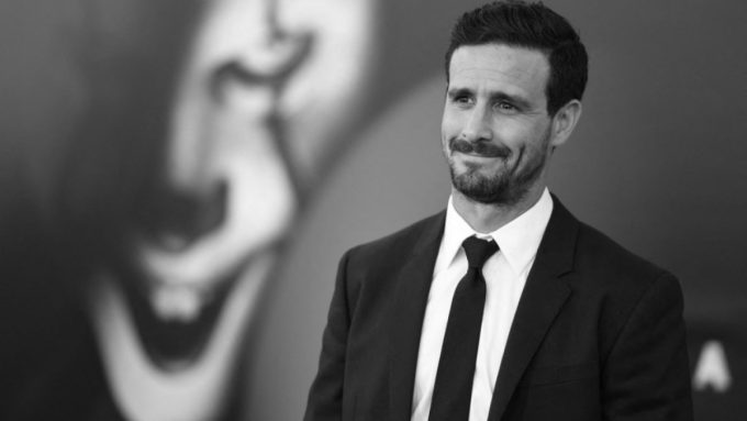 A black and white portrait of actor James Ransone smiling slightly while looking off-camera. He is wearing a dark suit, white shirt, and dark tie. In the blurred background, a large graphic of a clown face is partially visible.