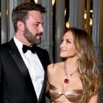 Jennifer Lopez Ben Affleck dressed in formal attire at an evening event, with Lopez wearing a gold dress and large necklace while looking up at Affleck in a tuxedo.