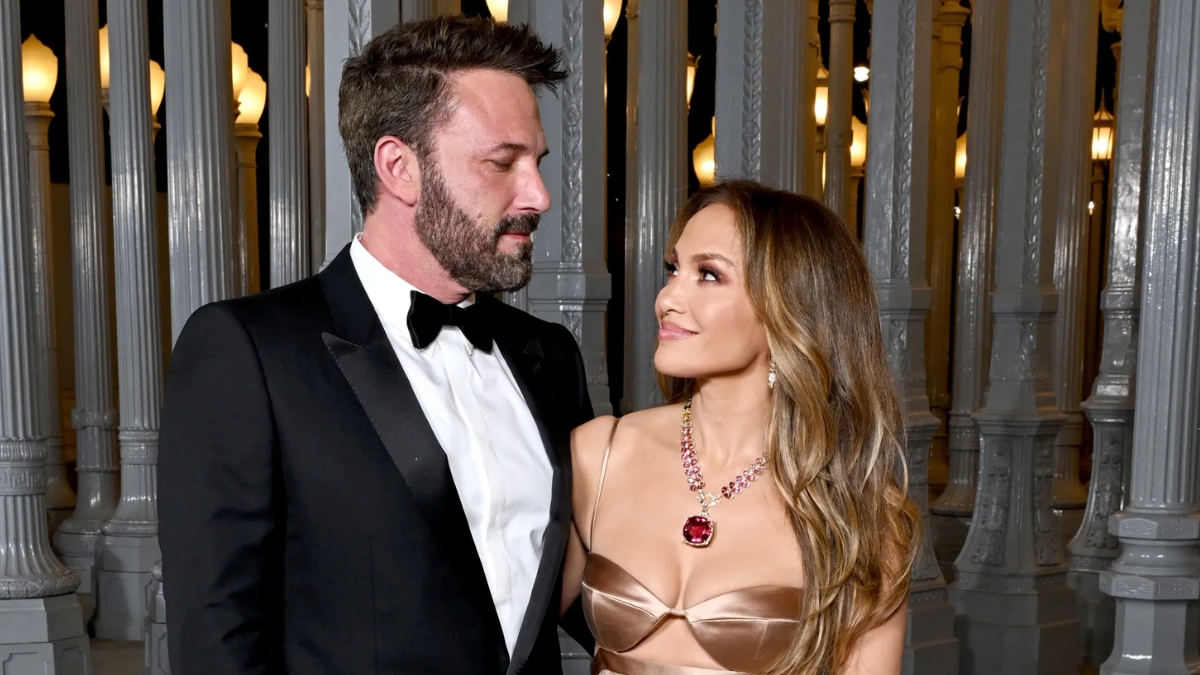 Jennifer Lopez Ben Affleck dressed in formal attire at an evening event, with Lopez wearing a gold dress and large necklace while looking up at Affleck in a tuxedo.