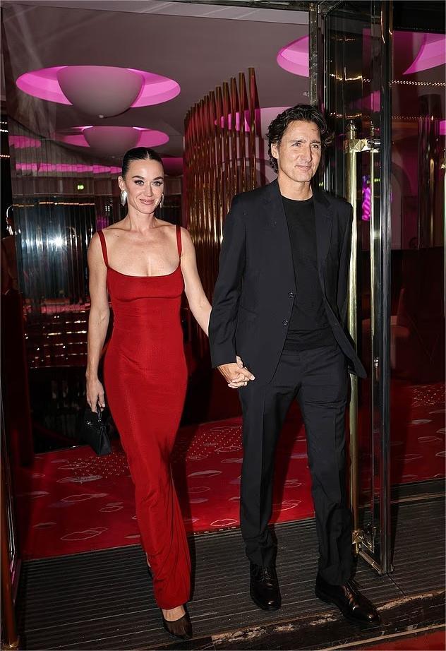 A full-length color photo of a woman in a long, form-fitting red dress, who appears to be Katy Perry, holding hands with a man in a black suit, who appears to be Justin Trudeau. They are walking together, looking toward the camera as they exit a doorway.