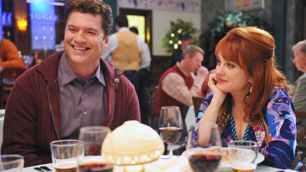 A medium shot of actor Pat Finn and guest star Wendi McLendon-Covey seated at a restaurant table. Pat, on the left, is wearing a maroon zip-up hoodie over a checkered shirt and is laughing heartily. Wendi, on the right, has red hair and is resting her chin on her hand, looking toward him with a slight smile.